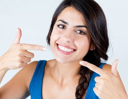 A young female with dark brown hair and a blue tank top pointing at her whiter, brighter teeth after seeing her cosmetic dentist