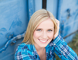 A young woman with blonde hair and a blue, plaid shirt smiles after receiving her veneers